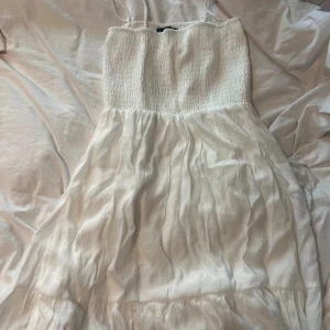 Vit sommarklänning med smock - Superfin vit klänning med smala axelband och smockat. Kjolen är luftig och har en lätt rynkad struktur som ger en flowy känsla. Perfekt för varma dagar och har en enkel men trendig look.