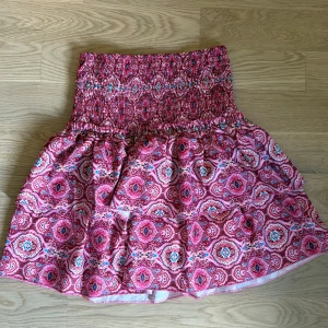 Rosa mönstrad kortkjol med smock - Supersöt kortkjol i rosa med färgglatt paisleymönster och bred smock i midjan. Kjolen har volang och är perfekt för dig som gillar färg och mönster. Lätt och luftig känsla, passar perfekt till varma dagar.