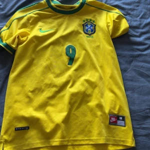 Brasilien Ronaldo 9 Nike fotbollströja - Gul Brasilien fotbollströja med gröna detaljer, nummer 9 och Ronaldo på ryggen. Klassisk Nike-logga på bröstet och landslagsemblem. Kortärmad modell i lätt och ventilerande material, perfekt för match eller träning.