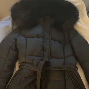 Svart lång pufferjacka med fuskpäls - Säljer en svart lång pufferjacka med stor fluffig fuskpäls på huvan och bälte i midjan. Jackan har quiltad design, dragkedja och knappar framtill. Perfekt för kalla dagar när du vill vara både varm och snygg. Passar xs och s