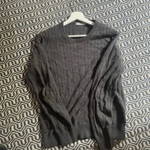 Mörkgrå kabelstickad tröja J.Lindeberg - Kabelstickad mörkgrå tröja från J.Lindeberg i klassisk modell med rund halsringning. Tröjan är långärmad och har ribbade muddar vid ärmslut och nederkant. Perfekt för dig som gillar stilrena och tidlösa plagg med snygg struktur.