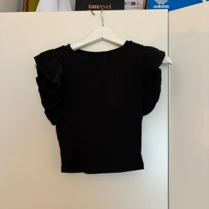 Svart topp med volangärmar - Säljer en svart topp med breda volangärmar som ger en cool och trendig look. Toppen är croppad och har en rund halsringning. Perfekt för dig som vill sticka ut lite extra med detaljerade ärmar.