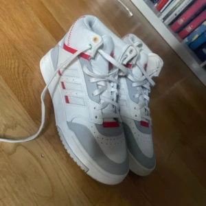 Adidas vita och röda sneakers high - Snygga vita Adidas sneakers med röda detaljer och högt skaft. Skorna har klassiska tre ränder på sidan, snörning och coola symboler på hälen. Ovandel i syntet och textil, Ortholite-innersula för extra komfort. Perfekta för dig som vill sticka ut med stil. Använda 4-5 gånger.