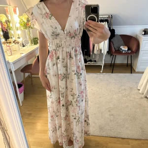 Blommig långklänning med v-ringning - Romantisk långklänning i ljus bas med blommigt mönster i rosa och grönt. Djup v-ringning fram och öppen rygg, resår i midjan och slits bak. Lätta, korta ärmar och luftig känsla – perfekt för varma dagar.
