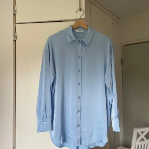 Ljusblå skjorta från Primark - Säljer en ljusblå skjorta från Primark med klassisk krage och knappar hela vägen framtill. Skjortan har lång ärm med manschett och är tillverkad i ett mjukt, lite glansigt material som ger en snygg look. Passformen är loose och skjortan är perfekt för en chill och stilren vibe.