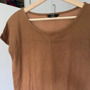 Velvet brun t-shirt  - En enkel brun t-shirt med rund halsringning och korta ärmar. Toppen har en rak passform och synliga sömmar framtill som ger en stilren look. Perfekt att matcha med jeans eller kjol för en avslappnad vibe.