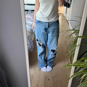 Blå ripped jeans från Zara - Säljer ett par ljusblå jeans från Zara med rejält slitna hål på båda benen. Modellen är loose fit med raka ben och klassisk femficksdesign. Jeansen har hög midja och är tillverkade i bomull. Perfekta för en avslappnad och trendig look.