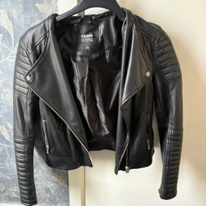 Svart skinnjacka från Cropp XS - Snygg svart skinnjacka från Cropp i bikerstil med coola detaljer som quiltade partier på axlar och ärmar samt dragkedjor vid fickor och framtill. Jackan har en klassisk krage och en slim passform som ger en edgy vibe.