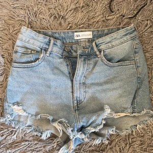 Ljusblå jeansshorts från Zara - Säljer ett par ljusblå jeansshorts från Zara i klassisk femficksmodell. Shortsen har råa fransiga kanter och slitna detaljer för en avslappnad vibe. Perfekta till sommaren och lätta att matcha med allt. Snyggt och trendigt plagg för dig som gillar denim!