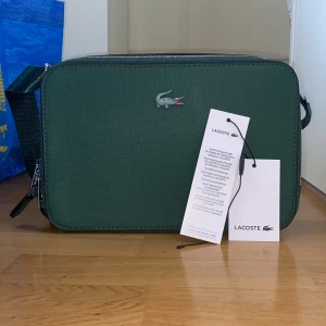 Grön axelväska från Lacoste - Snygg grön axelväska från Lacoste med klassisk krokodil-logga framtill. Väskan har en rektangulär form, justerbar axelrem och dubbla dragkedjor i silver. Tillverkad i tålig polyester med innerfack och smarta detaljer. Perfekt för att bära dina essentials med stil.