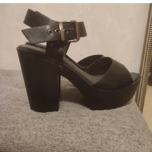 Svarta sandaler med klack och spänne - Snygga svarta sandaler med bred klack och platåsula. Skon har en bred rem över tårna och justerbart spänne runt ankeln. Perfekta för dig som vill ha både höjd och stabilitet. Materialet är troligtvis syntet.