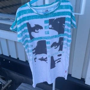 Randig t-shirt med tryck från Name It - Cool t-shirt från Name It i vitt med turkosa ränder och ett stort svartvitt ansiktstryck på framsidan. T-shirten har korta ärmar och är gjord i mjuk bomull, perfekt för en avslappnad stil. Passar dig som gillar grafiska prints och streetkänsla.