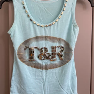 Ljusblå linnetopp med pärldetaljer - Ljusblå ärmlös linnetopp från T&R med coola pärldetaljer runt halsringningen och på det stora logotyptrycket framtill. Toppen är i mjuk bomulls- och polyestermix och har en slim passform. Perfekt för dig som gillar unika detaljer och en fräsch look.
