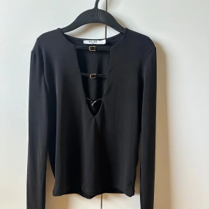 Svart långärmad topp från NA-KD - Svart långärmad topp från NA-KD med djup v-ringning och snygga dekorativa spännen framtill. Toppens minimalistiska design gör den lätt att matcha och den har en tight passform som framhäver siluetten. Perfekt för dig som gillar trendiga detaljer.