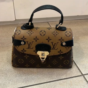 Brun handväska - Snygg handväska från . Väskan är brun och beige med svarta detaljer och guldfärgat spänne. Tillverkad i skinn med ett robust handtag och loggor över hela väskan. Perfekt för dig som vill sticka ut med en lyxig accessoar.
