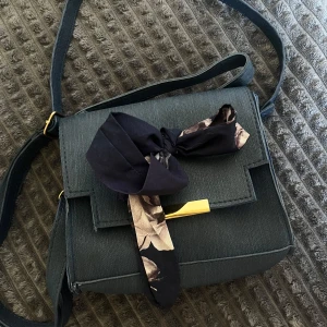 Svart axelväska med scarfdetalj - Stilren svart axelväska i tyg med guldiga metalldetaljer och en dekorativ scarf i svart och beige knuten framtill. Väskan har en fyrkantig form och ett unikt gult innerfoder med blommigt mönster. Perfekt för dig som vill ha en personlig touch på din outfit.