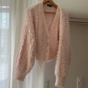 Ljusrosa fluffig kofta med knappar från missguided - Supermjuk och fluffig ljusrosa kofta från Missguided med v-ringning och två stora knappar framtill. Koftan är croppad och har långa ärmar med ett snyggt stickat mönster. Perfekt för lager-på-lager och en mysig vibe.