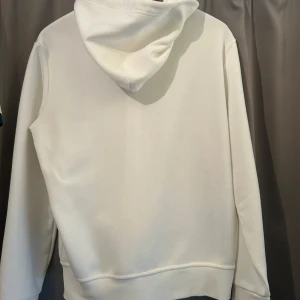 Vit hoodie från Polo Ralph Lauren M - Snygg vit hoodie från Polo Ralph Lauren i storlek M. Tröjan har dragkedja, snörning i huvan och klassisk logga på bröstet. Tillverkad i en mjuk mix av bomull och polyester, perfekt för chill dagar. Fickor framtill för extra stilpoäng.