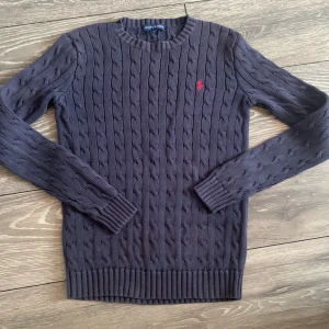 Mörkblå kabelstickad tröja Ralph Lauren - Kabelstickad mörkblå tröja från Ralph Lauren med klassisk rund hals och röd broderad logga på bröstet. Långärmad modell i mjuk bomull, perfekt för lager-på-lager och snygg till jeans eller kjol.