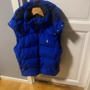 Blå dunväst från Polo Ralph Lauren - Ny pris 3899Snygg blå dunväst från Polo Ralph Lauren med klassisk broderad logga på bröstet. Västen har hög krage, dragkedja framtill och praktiska fickor med dragkedja. Perfekt för lager-på-lager och har en sportig vibe med sin quiltade design och huva.