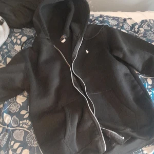 Svart hoodie från Polo Ralph Lauren - Svart hoodie från Polo Ralph Lauren med klassisk vit logga på bröstet. Tröjan har huva, dragkedja framtill och två stora fickor. Tillverkad i mjuk bomull, perfekt för chill dagar. Snygg och enkel att matcha med det mesta.