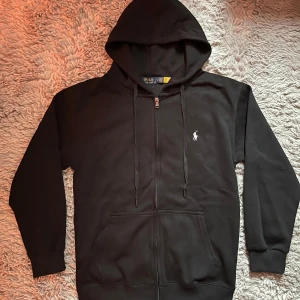Svart hoodie från Polo Ralph Lauren - Svart hoodie från Polo Ralph Lauren med dragkedja, huva och snörning. Klassisk vit logga broderad vid bröstet. Hoodien är helt ny och jag säljer den pga den inte passar mig. Hör av dig vid intresse👌
