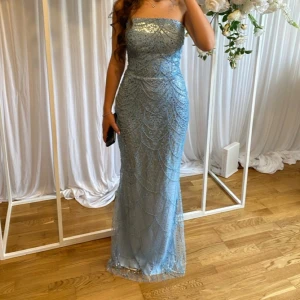 Ljusblå glittrig långklänning - Säljer en ljusblå axelbandslös långklänning med glittriga paljettdetaljer och snyggt mönster. Klänningen har figurnära passform och är perfekt för bal eller fest. Endast använd 1 gång. Skriv om ni har frågor💕