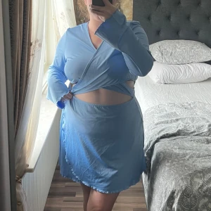 Ljusblå omlottklänning med cut-outs - Snygg ljusblå kort omlottklänning med långa ärmar och trendiga cut-outs i midjan. Klänningen har knyt i sidan och v-ringad hals, samt mjukt, stretchigt material som ger en bekväm passform. Perfekt för dig som gillar en unik och modern stil. Använd en gång. 