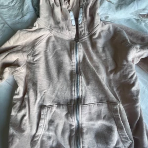 Beige hoodie med dragkedja - Mysig beige hoodie från H&M med dragkedja framtill och två stora fickor. Tröjan har huva med snörning och långa ärmar. Perfekt för chill dagar eller när du vill ha en enkel och stilren look.