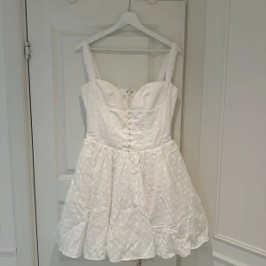 Vit broderad kortklänning House of CB - Supersöt vit kortklänning från House of CB med broderade blommor, knappar framtill och fluffig kjol med tyll under. Klänningen har breda axelband och dragkedja bak. Perfekt för sommarens alla tillfällen!