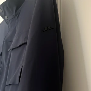 Navy rock från J.Lindeberg - Stilren navy rock från J.Lindeberg med hög krage och fyra stora fickor framtill. Jackan har diskret JL-logo på ärmen och stängs med knappar. Perfekt för dig som gillar minimalistisk och clean look. Pris kan även diskuteras 