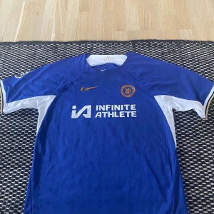 Chelsea Nkunku 18 matchtröja Nike - Chelsea FC matchtröja med Nkunku 18 på ryggen. Klassisk blå färg med vita detaljer på sidorna och ärmarna, orange Nike-logga och Chelsea-märke på bröstet. Materialet är lätt och andas, perfekt för fotboll. Sponsorlogga 'Infinite Athlete' på framsidan. Pris går att diskutera. 