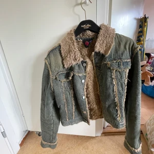 Y2k vintage Jeansjacka - Säljer en grå jeansjacka med mysigt beige teddyfoder på insidan och kragen. Jackan har råa, slitna kanter och knappar framtill. Perfekt för dig som gillar en cool och avslappnad stil med extra värme. Långärmad och med klassiska fickor framtill.