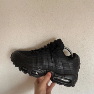 Svarta Nike Air Max 95 - Säljer ett par helsvarta Nike Air Max sneakers med chunky sula och synliga luftbubblor. Skorna har snörning, rund tå och är tillverkade i en mix av skinn och mesh för en sportig vibe. Perfekta för dig som gillar streetstyle och vill ha en clean look.
