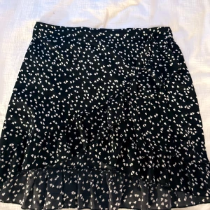 Svart kortkjol med vita hjärtan - Supersöt svart kortkjol med vita hjärtan och volangkant. Kjolen har omlottmodell och är perfekt för dig som gillar lekfulla detaljer. Lätt och luftig, passar perfekt till sneakers eller boots.