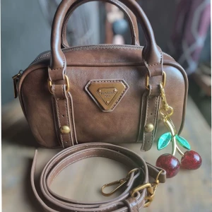 Retro brun handväska  - Retro brun handväska i pillow shape med handtag och avtagbar axelrem. Charmig körsbärsnyckelring ingår. Tillverkad i material av hög kvalitet PU läder som ger en känsla av äkta skinn. Endast använd en gång – i mycket gott skick. Mått: 12 x 21 x 8 cm. Axelrem är ungefär 95cm långt 