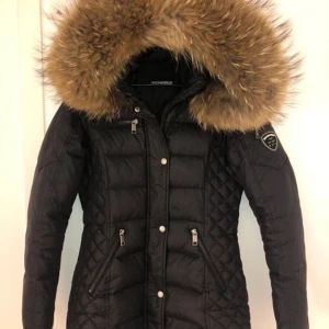 Rockandblue svart dunjacka med äkta päls  - Snygg svart dunjacka med stor fluffig äkta päls på huvan och quiltade detaljer på sidorna. Jackan har dragkedja, knappar och två snedställda fickor med dragkedja. Märkespatch på ärmen ger en cool touch. Perfekt för kalla vinterdagar. Köpt för 3799kr, säljes för 1700kr men pris kan diskuteras. 