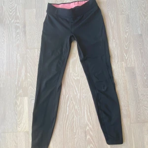 Svarta tights med snörning - Svarta träningstights med snörning i midjan och praktisk dragkedjeficka bak. Benen har reflexdetaljer, perfekta både för löpning och gym. Defekt förekommer på vänstra benet, se bild 5.