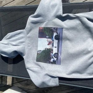 Grå Hardies NYC hoodie med biltryck - Säljer en grå hoodie från Hardies NYC med stor färgglad print på ryggen av en sportbil och folk på gatan. Framsidan har Hardies NYC-logga med en orange hund. Mjuk insida och klassisk känguruficka. Perfekt för dig som gillar streetwear och unika motiv.