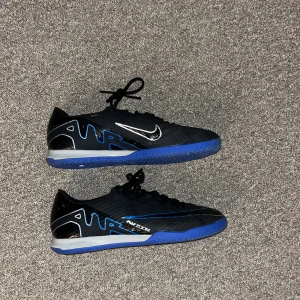 Nike Mercurial fotbollsskor svart/blå - Säljer ett par Nike Mercurial fotbollsskor i svart med blå och silverdetaljer. Skorna har snörning, låg profil och en blå platt sula med Air Zoom-dämpning. Perfekta för inomhusspel och har en sportig look med tydlig Nike-logga på sidan.