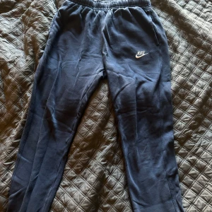 Mörkblå mjukisbyxor från Nike - Mörkblå mjukisbyxor från Nike med broderad vit logga på låret. Byxorna har resår i midjan och är tillverkade i mjuk bomullsmix. Perfekta för chill eller träning, med klassisk och enkel design.