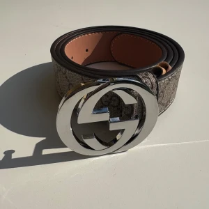 Gucci Bälte - Snyggt Gucci-bälte med klassiskt GG Supreme-mönster i beige och brunt. Bältet har en bred design och en stor, rund spänne i silver med dubbla G-loggan. Tillverkat i skinn med canvas-yttersida och stilren form – perfekt för att lyfta vilken outfit som helst.