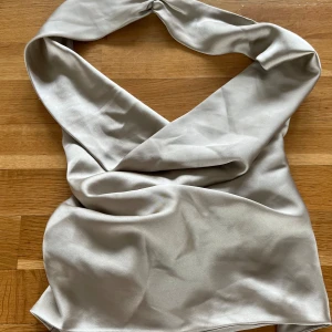 Champagnefärgad halterneck i satin - Supersnygg champagnefärgad halternecktopp i glansig satin med breda axelband och knyt i ryggen. Toppen har en draperad look framtill och är perfekt för dig som vill sticka ut med en elegant vibe. Passar till både jeans och kjol för en trendig stil.