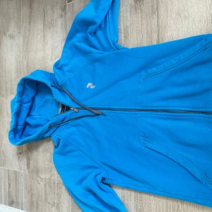 Blå hoodie från Peak Performance - Snygg blå hoodie från Peak Performance med dragkedja och huva. Tröjan har broderad logga på bröstet, två fickor framtill och är tillverkad i mjuk bomull. Perfekt för dig som gillar en sportig och avslappnad stil.