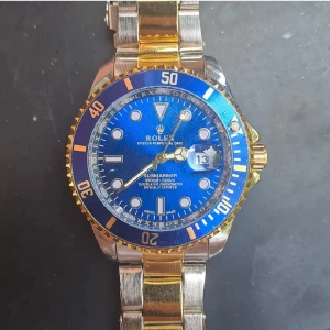 Klocka Submariner blå/guld herrklocka - Snygg  Submariner med blå urtavla och gulddetaljer. Klockan har en tvåfärgad länk i silver och guld, tydliga index och datumfönster. Klassisk rund boett och vridbar bezel i blått och guld. Perfekt för dig som gillar exklusiv stil och ikonisk design.
