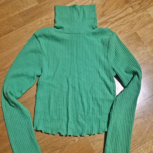 Grön ribbad polotröja från FB Sister - Snygg grön ribbad polotröja från FB Sister i storlek S. Tröjan har lång ärm, hög polokrage och är tillverkad i mjukt stretchigt material. Perfekt för lager-på-lager och ger en fräsch färgklick till din outfit.