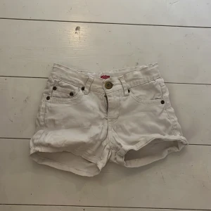 Vita jeansshorts med broderad krona - Snygga vita jeansshorts från DXEL med klassisk femficksdesign och coola broderade kronor på bakfickorna. Shortsen har uppvikta benslut, bälteshällor och stängs med dragkedja och knapp. Perfekta för sommardagar när du vill sticka ut lite extra.
