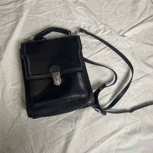 Svart axelväska i skinn från The Trend - Snygg svart axelväska från The Trend, tillverkad i äkta skinn. Väskan har ett klassiskt rakt format lock med silverfärgat lås och både handtag och justerbar axelrem. Flera fack inuti för smidig organisering. Perfekt för dig som gillar stilren och tidlös design.