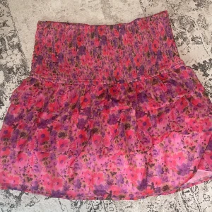 Blommig volangkjol från BikBok - Supersöt kort kjol från BikBok med blommigt mönster i rosa, lila och röda toner. Kjolen har bred smock i midjan och flera volanger för extra flowy känsla. Perfekt för dig som gillar färg och vill sticka ut lite extra!