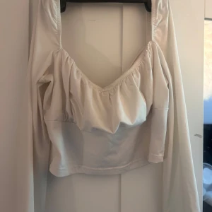 Vit croppad topp från SHEIN - Supersöt vit croppad topp från SHEIN med långa ärmar och snygg v-ringning. Toppen har rynkade detaljer över bysten och är i ett mjukt, stretchigt material. Perfekt för dig som gillar trendiga och feminina plagg med lite extra volym upptill.